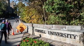 Students walk through campus on a sidewalk next to a long granite sign that says Cornell University 
