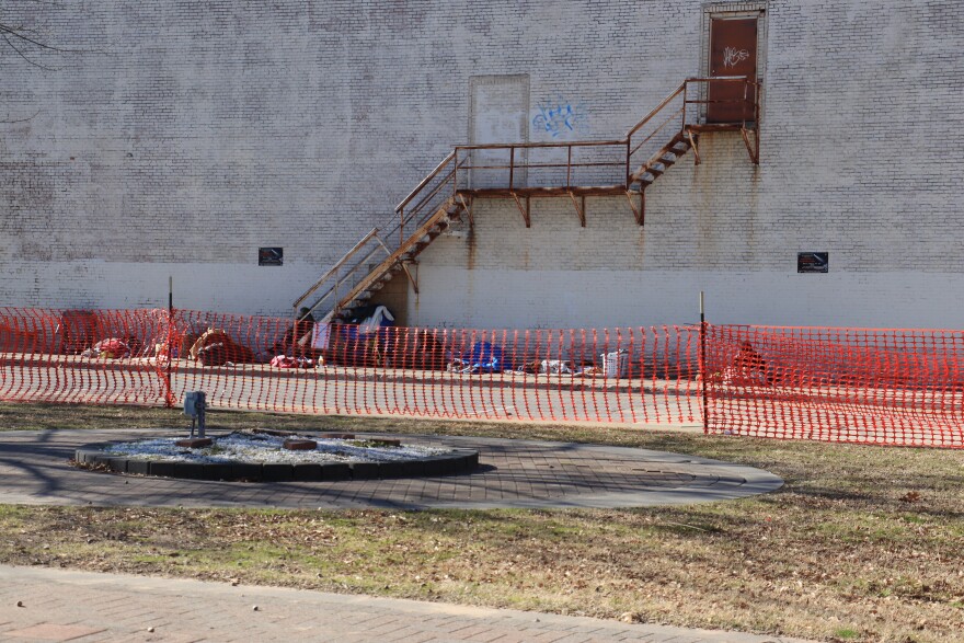 People sit on the sidewalk across the street from Celebration of Life Park, which is surrounded by a temporary fence and "no trespassing" signs.