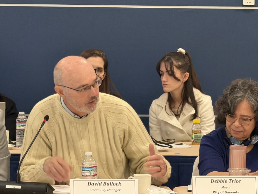 A man in a cream colored sweater speaks at a government meeting