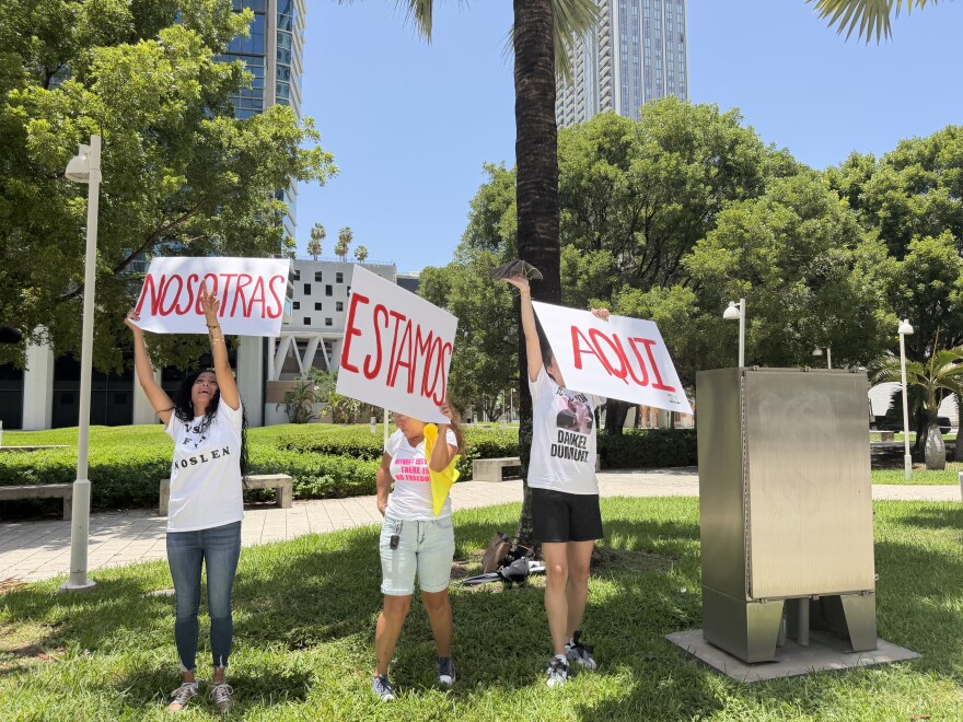 Anisley Cortes, Judith Castellano and Anett Uset-Dumont wave signs that read "NOSOTRAS ESTAMOS AQUI" (We are here) towards the windows of FDC Miami on July 15, 2026.