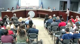 A school board meeting, from behind the seated audience, facing trustees and a speaker