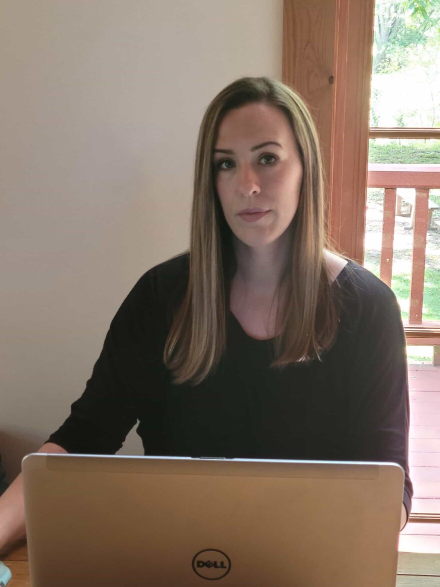 Jeanna Norris stands in front of a gray Dell laptop computer. She has light brown hair with blond streaks and is wearing a long sleeve black shirt. A porch can be seen on the background. 