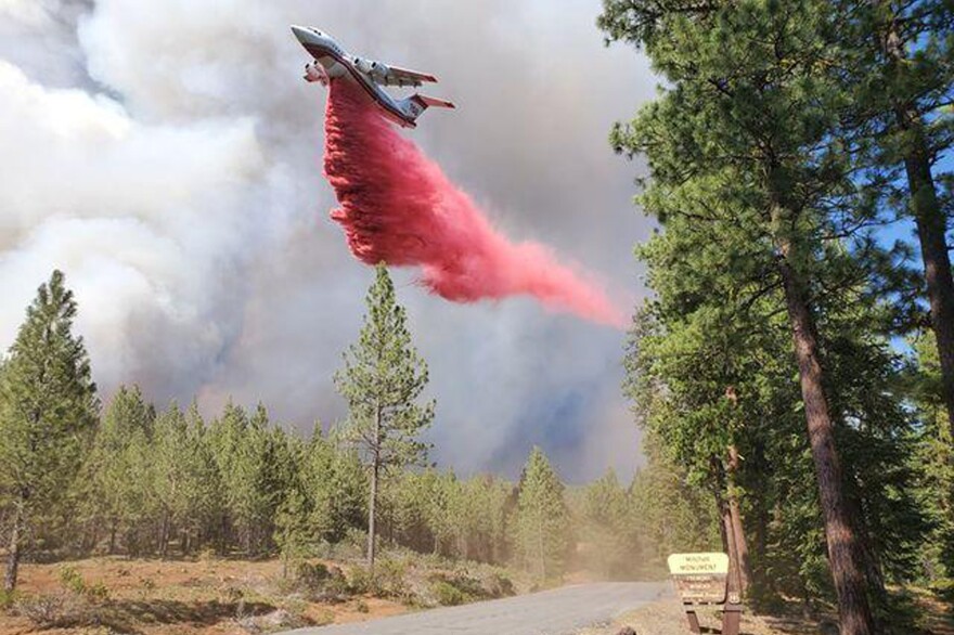 A plane drops red fire retardant over a wooded area. The air is filled with smoky haze and barely any blue sky can be seen.