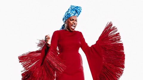 A woman in a dramatic, tassled. floor-length red dress and blue head turban, singing forcefully