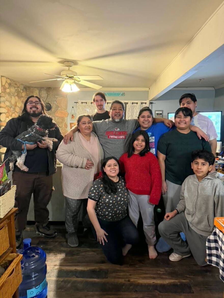 Ten people smiling in a group family photo. A middle-aged man is in the middle, with his arms outstretched around the two people on either side of him. A younger woman, his daughter, is to his right and an older woman, his wife, is to his left.