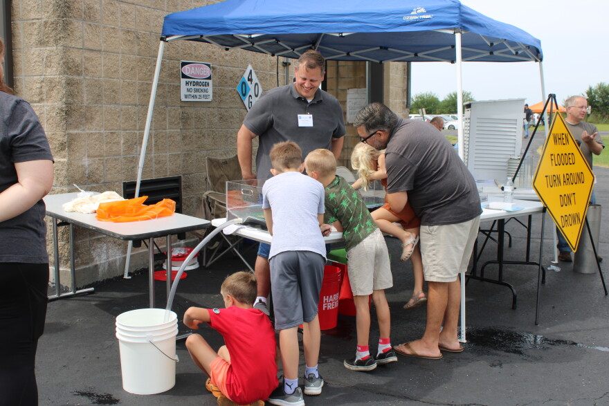 A man stands in front of a table holding a small girl up so she can reach. Other children crowd around the table while a National Weather Service employee looks on