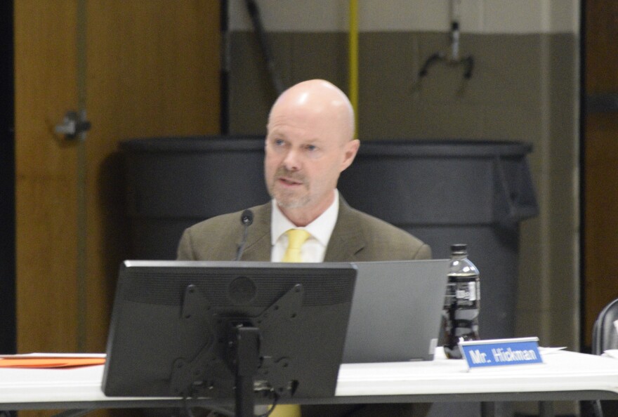 Man in suit with yellow tie speaks. In front of him is a nametag and laptop.