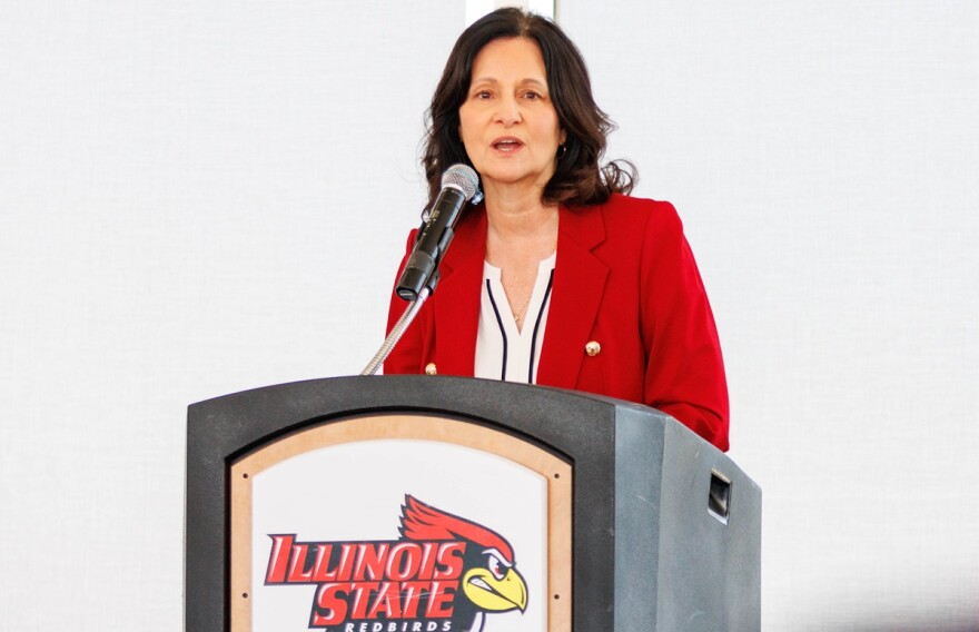 Jeri Beggs standing at a podium wearing a red jacket and white blouse. The podium has the Illinois State Redbird logo that features a red and gold-colored animated redbird