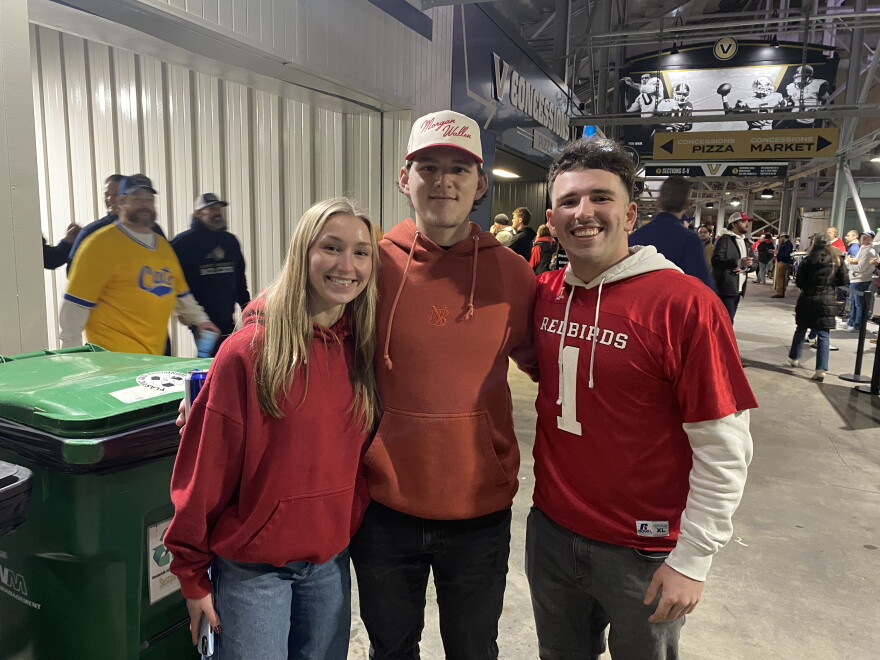 Three young adults, two men and one woman, smile and pose together indoors at a stadium. All are wearing red tops, with the man on the right in a "Redbirds" jersey. Other people and concession stands are visible in the background.