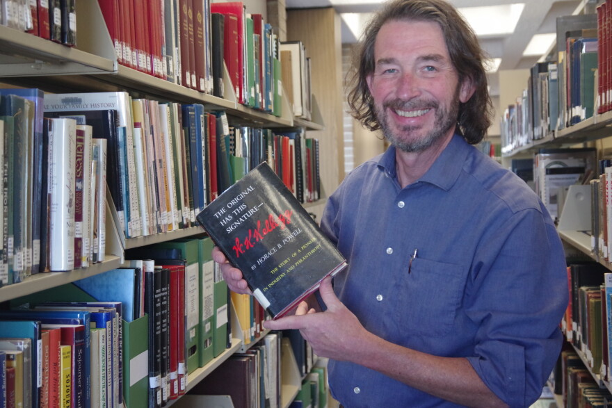 A man in a long sleeve blue button up holds up a black book as he smiles. He stands between two shelves filled with books inside a public library.