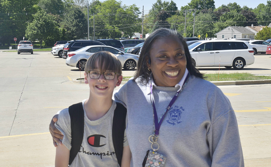 A woman smiles as she stands with a boy in a backpack, with parking spaces behind.