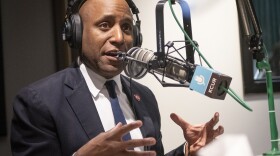 A man wearing a blue suit and tie sits inside a radio studio. He is talking at a microphone and gesturing with both hands.
