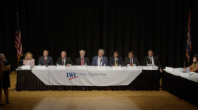 Seven men and a woman sit at along table. They're flanked on the stage by a table of a few journalists and a woman standing while speaking into a microphone. The ensemble looks a bit like "The Last Supper."