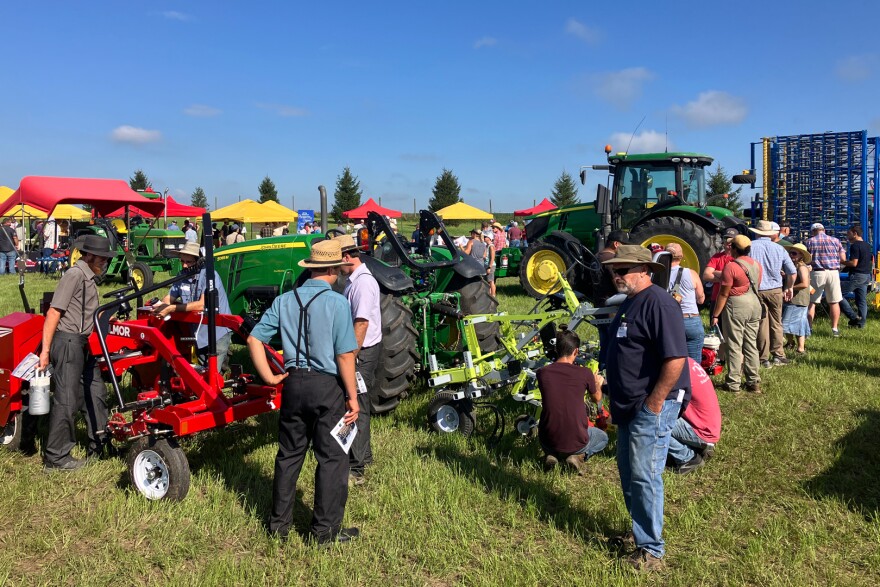 People in hats and long pants cluster around a line of small- and large-scale equipment in a grassy field.