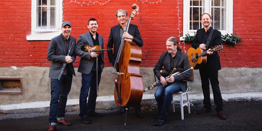 Five men standing in front of a red brick wall holding string and woodwind instruments.