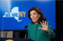 Gov. Kathy Hochul with her hand raised, wearing green, in front of a "WE ARE NY" monitor.