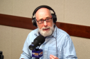 A bald man with a bushy grey beard is sitting in front of a microphone at a table in a radio talk studio and wearing eyeglasses and a light blue button-down shirt.