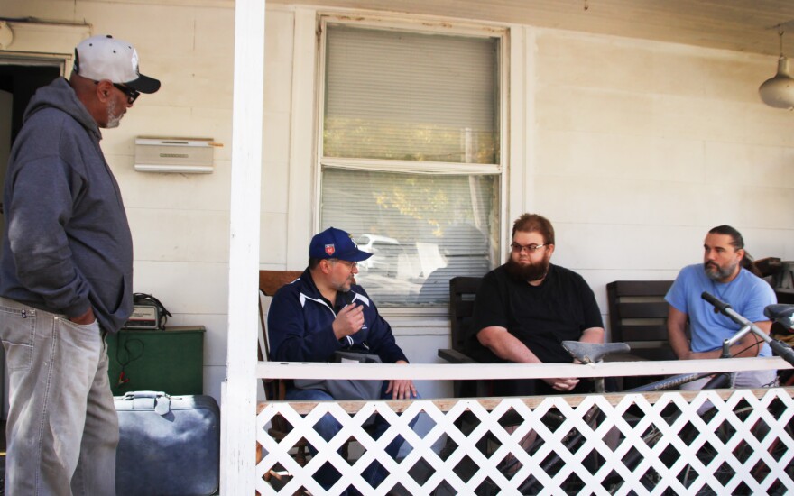 One man stands at left while three others sit in chairs on the front porch of a wood-framed, white house. The man seated third from left is talking.