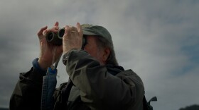 A man with gray hair is wearing an olive green baseball hat and utilitarian coat. He holds olive green binoculars to his eyes and looks through them at a three-quarter angle.