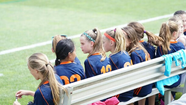 A girls' soccer team sits on a bench on the sideline.
