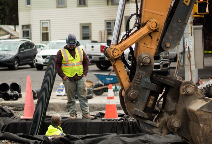 Two construction workers talk next to a piece of heavy machinery. There is a large whole one of the workers is in, with cones surrounding it.