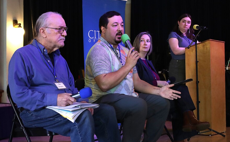  A group of people sit on a stage, one person speaks into a microphone, one person is at a podium.