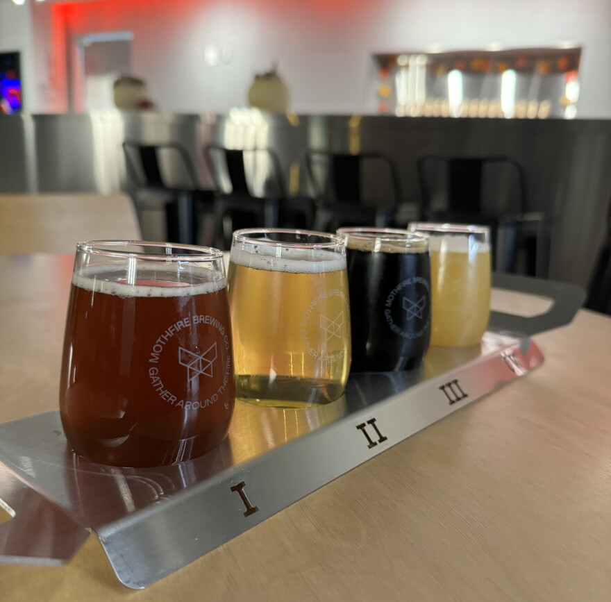 Four small glasses of craft beer arranged in a numbered metal flight tray on a wooden table at Mothfire Brewing Company.