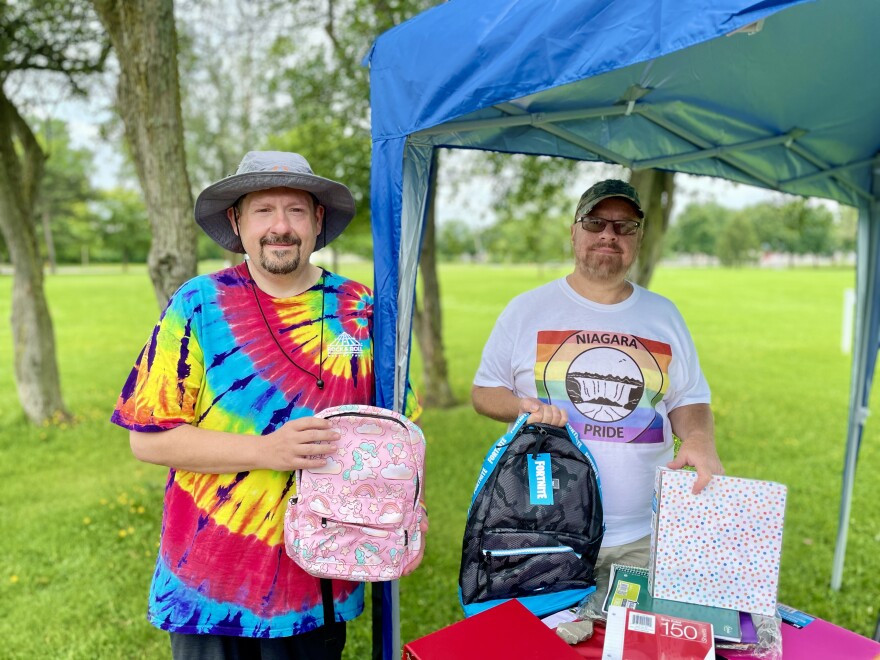 Two men stand under a gazebo tent holding backpacks and a binder. Ronald Piaseczny, on the left, is wearing a grey bucket hat and rainbow tie-dye shirt. Mark Smith, on the right, is wearing a baseball cap, glasses, and a shirt with the Niagara Pride logo on it.