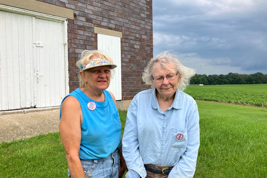 Two women in blue shirts stand in front of a barn with a soybean field and storm clouds in the distance.
