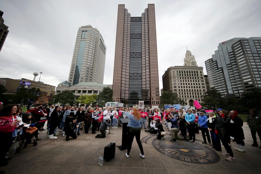 a woman stands at a podium facing a group of people gathered around her with the Columbus, Ohio, skyline in the background