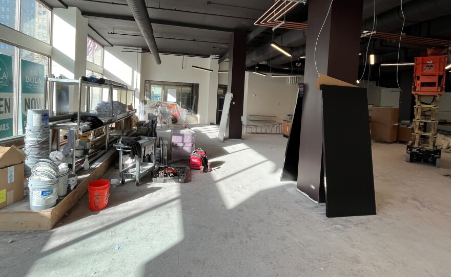 Buckets and other materials are stacked on a low-rise stage near the East Main Street windows of Sibley Square. The windows are covered in placards that read "La Bola, Open, Cocktails." The interior of the future space -- to be called The Duke -- is largely empty. A lift sits off to one side, raised nearly to the ceiling, which is painted black. Exterior walls are white.