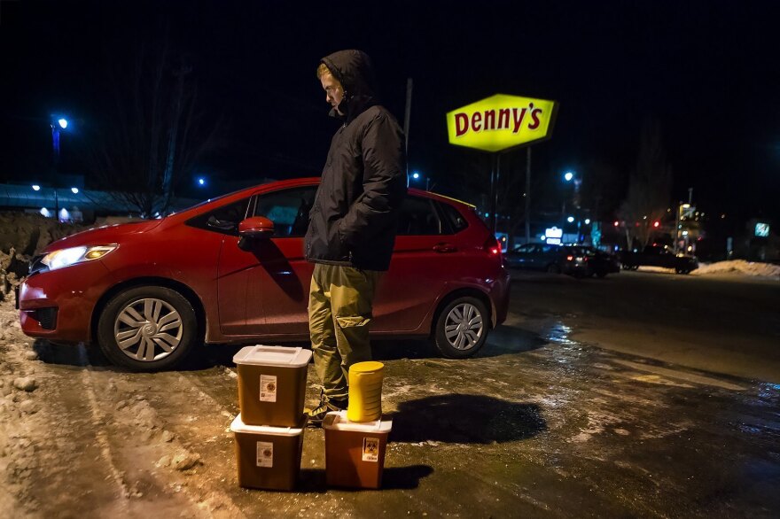 Jesse Harvey, founder of the Church of Safe Injection, stands in a Denny's parking lot in Auburn, Maine, alongside four sharps containers filled with used needles collected from drug users around the neighboring city of Lewiston.