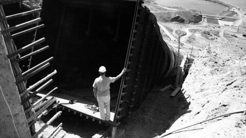 A man in a hard hat stands inside a massive dry pipe, looking down to a reservoir downhill 