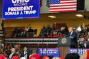 President Donald Trump speaks at a podium in a high school stadium full of supporters, with an american flag behind him