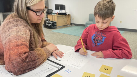 An adult in a sweater and glasses watches a child in a red hoodie complete a worksheet. 