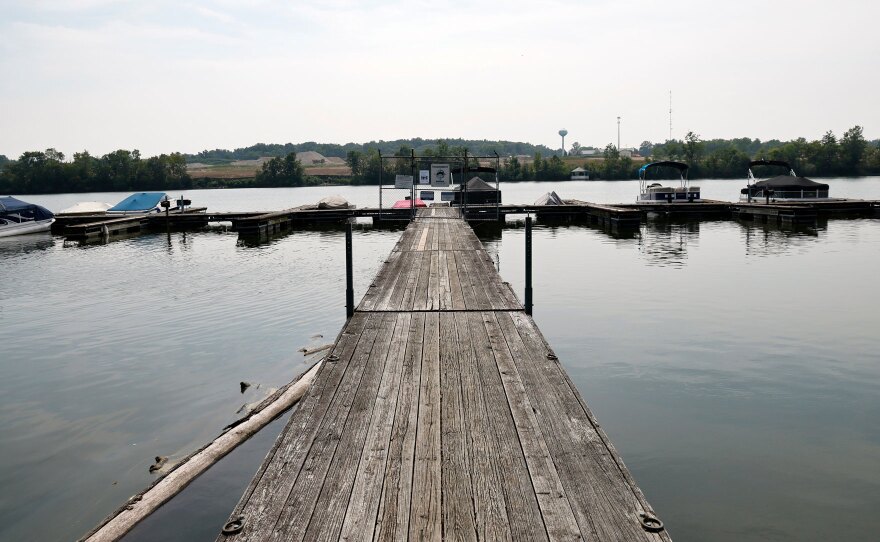 A wooden pier extends into a calm lake with boats docked on either side. The shoreline is lined with trees, and there are gentle hills in the background under a partly cloudy sky.