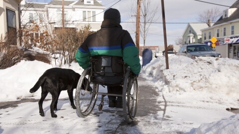 A stock photo of a woman with multiple sclerosis using a wheelchair, accompanied by a service dog. She is wheeling down a sidewalk surrounded by snow.