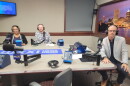 Three people wearing headphones sit at a table in a radio talk studio: A woman back left has her dark hair pulled back. She is wearing glasses and a dark top under a black sweater with a white border; a man back center has short red hair and is wearing glasses and a blue and white plaid button-down shirt; a man front right has very short dark hair and is wearing glasses, a black button-down shirt under a grey suitcoat, jeans, and brown shoes.