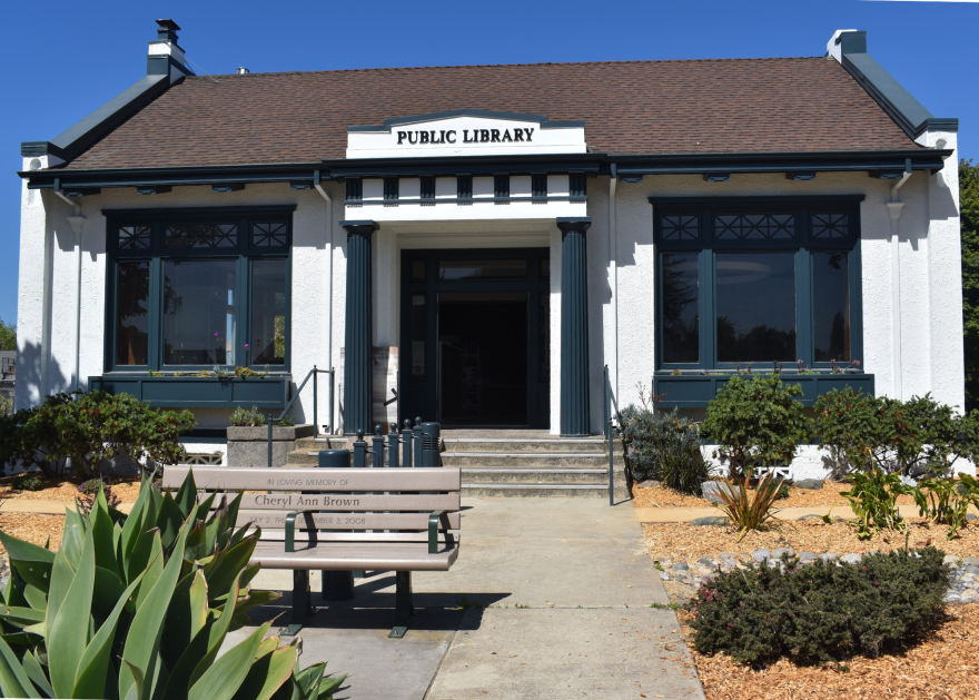 A small one-story building has the words "Public Library" over the door, steps up to the front door and a park bench in the foreground. 