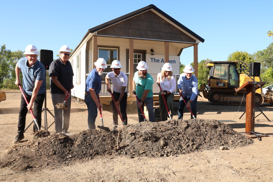 Members of Watered Gardens Ministries break ground at Freedom Village on September 4. They're wearing hard hats and are holding shovels in front of a pile of dirt/ They're standing in front of a model tiny home.