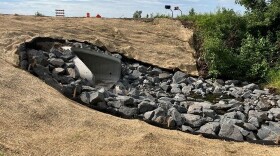  This recently completed 88” arch centerline culvert improves drainage beneath Hwy 169 near 435th Ln. Shows a large drainage pipe flowing into some large gray rocks underneath the roadway.