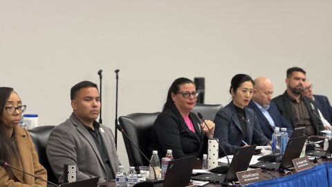 Seven councilmembers sit at a table in front of computers and microphones 