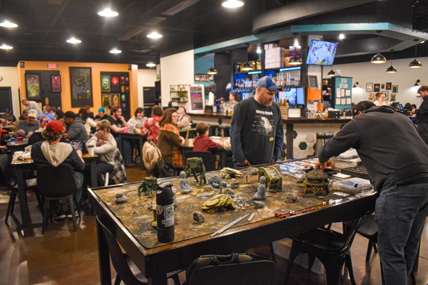 Two men lean over a large table covered in small set pieces and miniatures in a crowded café.