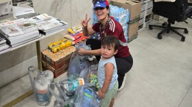 A woman and a young boy are next to grey plastic bags filled with supplies like paper towels.