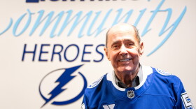 Older bald man wearing blue hockey jersey and smiling into the camera with a logo of a lightning bolt behind his right shoulder