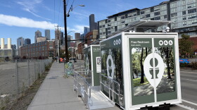 A photo of two Throne bathrooms with the Seattle skyline visible in the background. The bathroom trailers have images of trees on the outside and signage that says "free restroom" and an image of a toilet. 