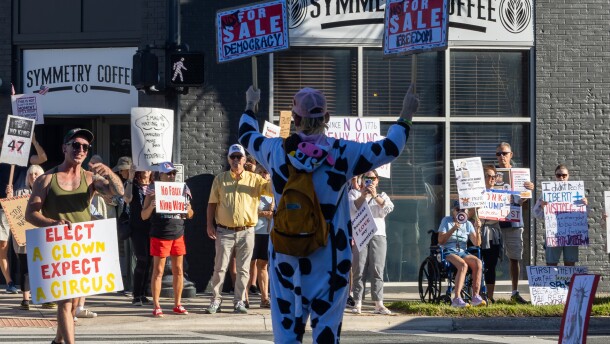 A man in a cow costume stands on a cross walk in downtown Ocala holding signs that say, "Not For Sale Democracy" and "Not For Sale Freedom" on Saturday, Oct. 18, 2025. (Gabriel Velasquez Neira/WUFT News)