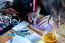 A man with long brown hair leans down close to a wooden table inside a brewery to write in Sharpie marker across a small zine made from purple construction paper. The table is littered with art supplies.