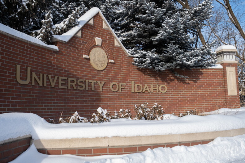 A red brick structure with the University of Idaho on it in gold letters. There is a planter in front with dead plants and snow in it. 