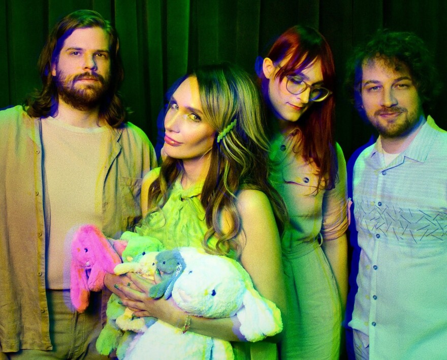 A woman holds four toy rabbits and looks at the camera with a wry expression while two men and another woman stand alongside her.